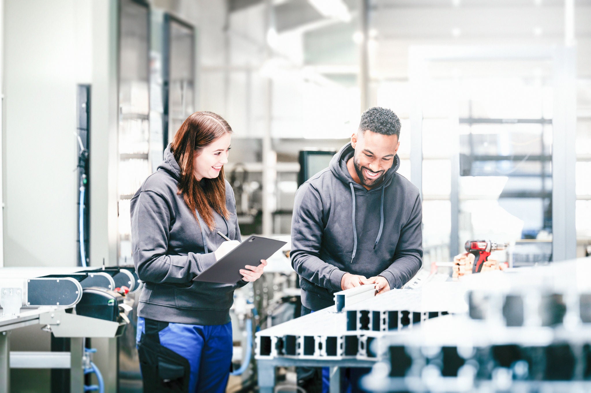 Ein Metallbau-Arbeiter bearbeitet in der Werkstatt Aluminumprofile. Eine Metallbau-Arbeiterin steht mit einem Zettel-Klemmbrett daneben.